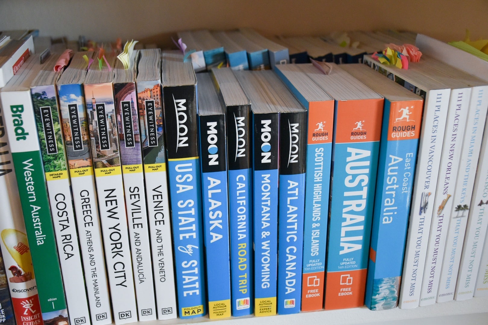 Bookshelf full of travel inspiration books, some with post it notes inside