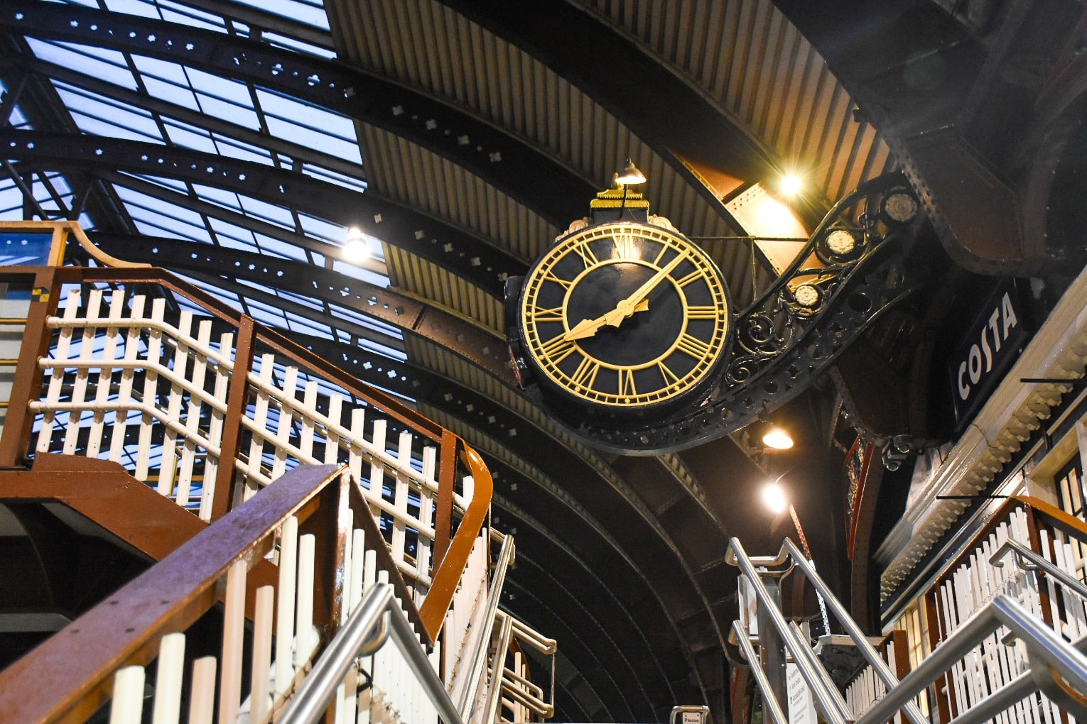 Photo of a black Victorian clock face with gold numerals and hands, positioned over the white and brown railings of a train station stairway. Metal arches across the roof